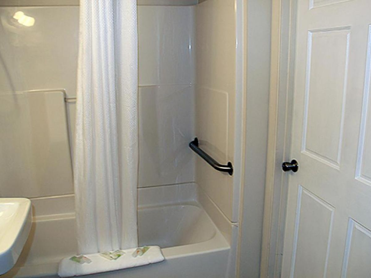 The image shows a bathroom with a tub, a shower curtain, a sink, a grab bar, and a closed door. The atmosphere appears clean and tidy.