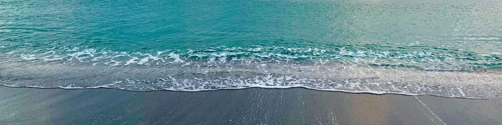 A serene beach scene with gentle waves lapping at the shore and clear blue-green water stretching into the horizon under a calm sky.