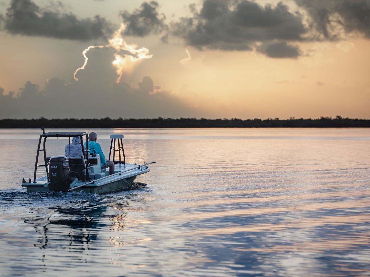 A small boat with two people is floating on calm waters during a picturesque sunset, with dramatic clouds in the background.