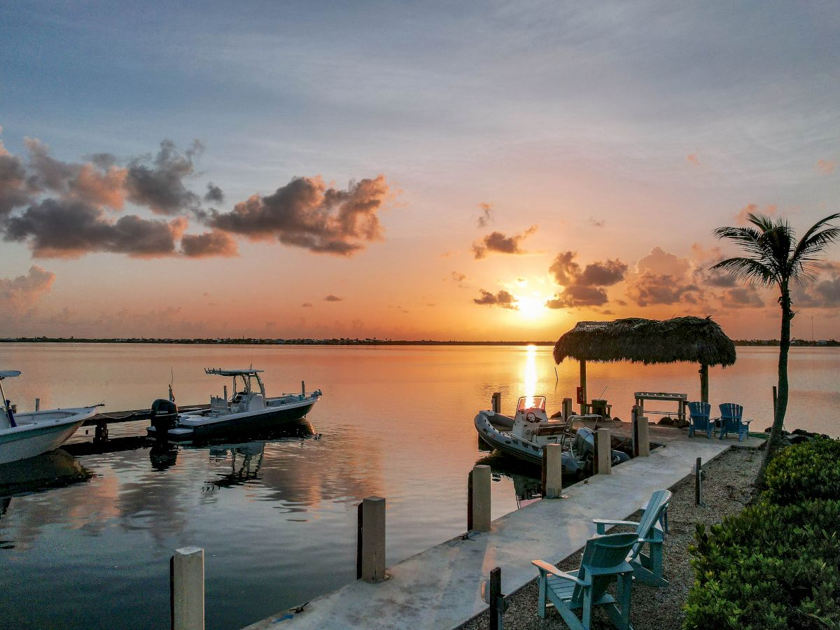 A serene sunset over a calm water body with boats docked, a palm tree, and a thatched shade.