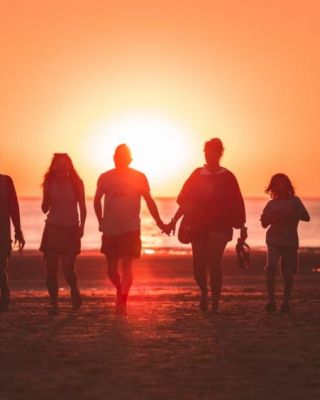 A group of five people is walking on the beach at sunset, silhouetted against the orange sky while holding hands.