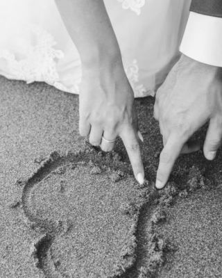 A bride and groom draw a heart in the sand with their fingers, celebrating their wedding in a romantic beach setting, showcasing their love.