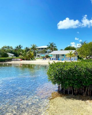 This image shows a peaceful waterfront scene with clear water, lush greenery, and charming houses under a bright blue sky with a few clouds.