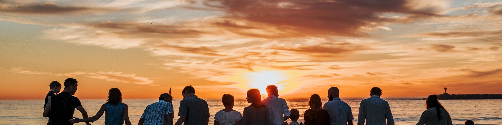 A group of people holding hands stands on a beach, facing a sunset over the ocean, silhouetted against a colorful sky with scattered clouds.