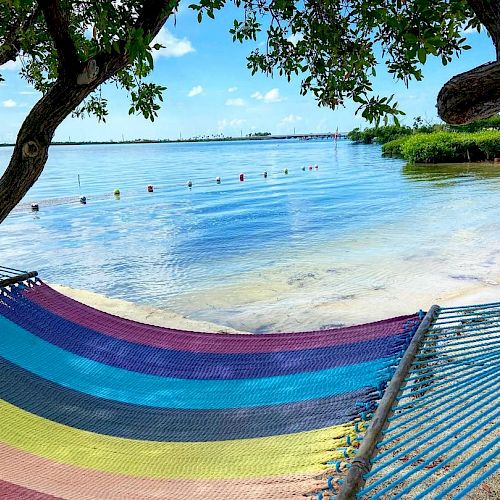 A colorful hammock is hung between two trees by a serene beach with clear blue water, distant shoreline, and a partly cloudy sky in the background.