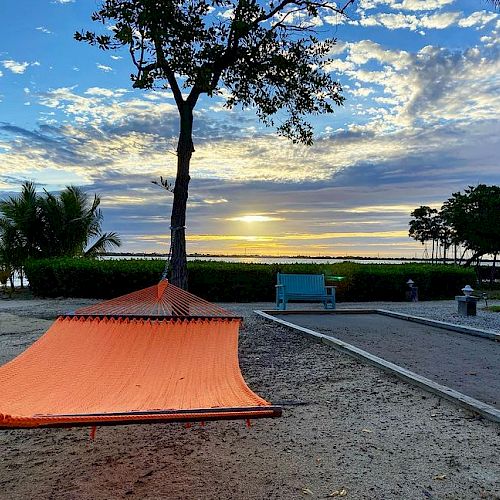 An orange hammock tied to a tree, a blue bench, and a serene beach with a beautiful sunset in the background.