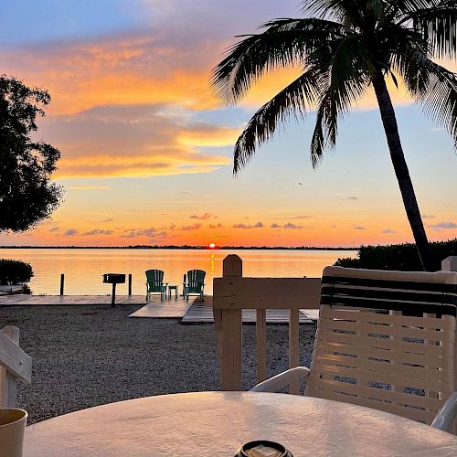 The image shows a tranquil beach scene at sunset, with palm trees, a table and chairs, and the sun setting over the water in the distance.