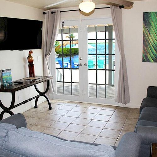 A cozy living room with gray couches, a wall-mounted TV, a decorative table, and a picture. French doors open to a patio with a pool view.