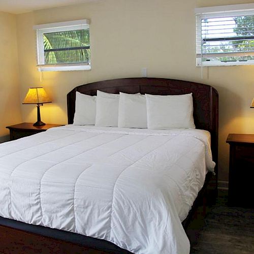 A well-lit bedroom with a large bed, white bedding, two bedside tables with lamps, a painting on the wall, and two small windows with blinds.