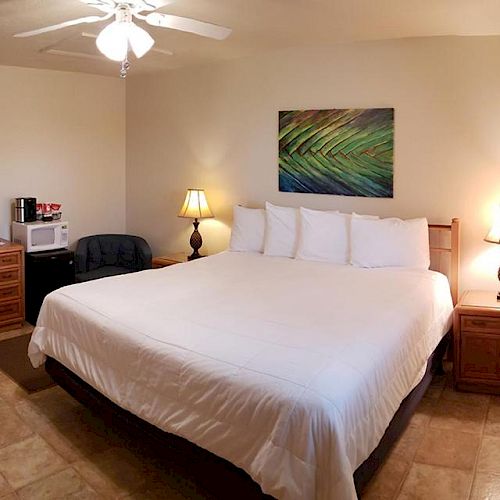 The image shows a neatly arranged hotel room with a large bed, bedside tables, lamps, a dresser with a mirror, and a small kitchenette.