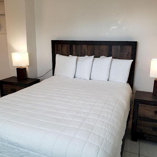 The image shows a bedroom with a bed, white bedding, wooden headboard, and two bedside tables, each with a lamp. The room has a door to the left.