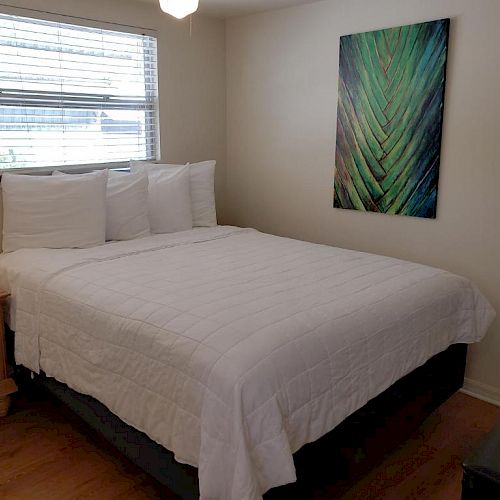 A small bedroom featuring a neatly made bed with white linens, a bedside table, a window with blinds, and a colorful abstract wall painting.