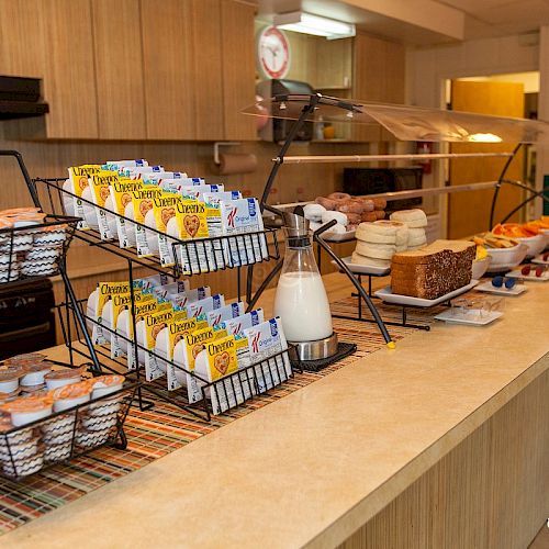 The image shows a breakfast buffet with cartons of juice, milk, various breads, muffins, fruit, and condiments on a counter.