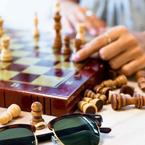 A person is moving a chess piece on an upright chessboard, with scattered pieces and a pair of sunglasses on the table.