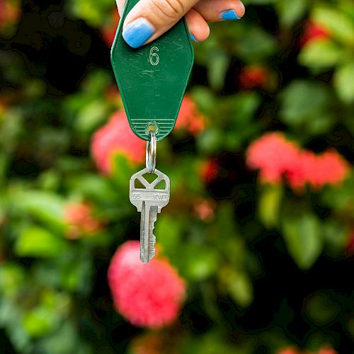 A hand with blue nail polish holds a key attached to a green keychain labeled '6,' with a background of blurred leaves and red flowers.