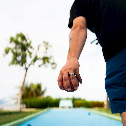 A person is playing bocce ball on a blue court, aiming to throw a ball forward with trees and greenery in the background.