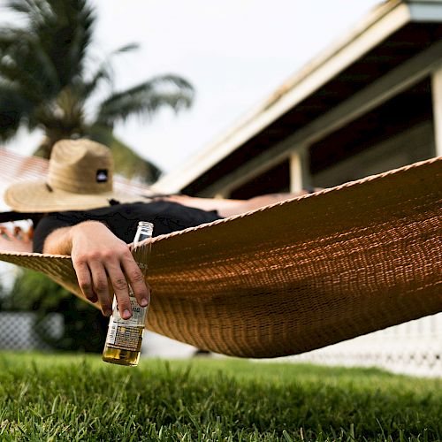 A person lies in a hammock, wearing a hat and holding a bottle, with a house and palm trees in the background.