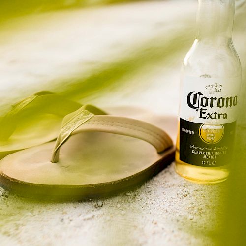 The image shows a pair of flip-flops and an open bottle of Corona Extra beer placed on a sandy surface, with green leaves partially framing the view.