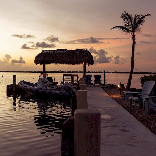 A serene dock at sunset with a thatched-roof hut, a boat tied to the side, a palm tree, and a few dockside chairs by the water.