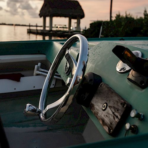 A close-up of a boat's steering wheel at sunset, with a dock and hut in the background across the water.