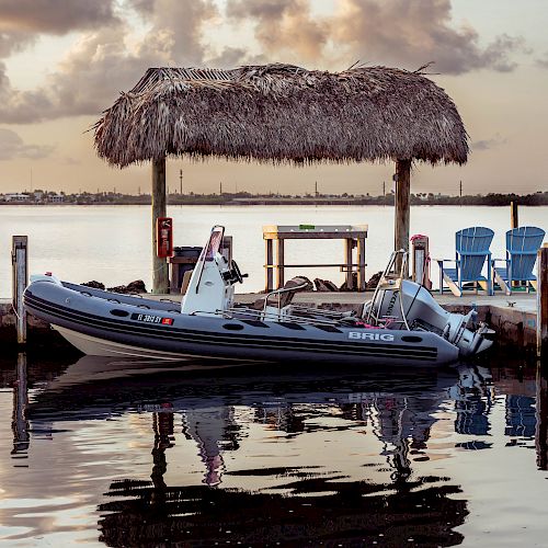 An inflatable boat is docked next to a pier with a thatched roof structure and blue chairs under a cloudy sky reflecting on calm water.