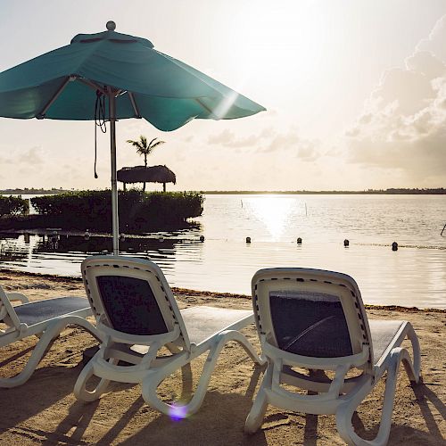 Two lounge chairs under a blue umbrella face a serene water body, with the sun setting on the horizon, casting shadows on the sandy shore.