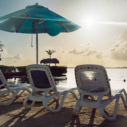 White lounge chairs and umbrellas on a sandy beach facing a calm body of water with a setting sun in the background.