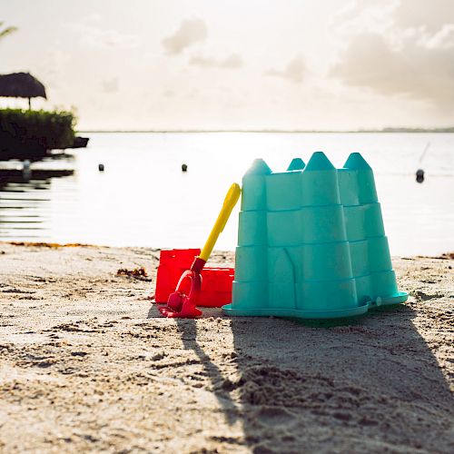 A plastic sandcastle mold and shovel are placed on a beach, with a calm body of water and a small hut in the background.