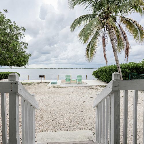 A beach view with a palm tree, two green chairs facing the sea, and a grill near the water, seen from a wooden deck.