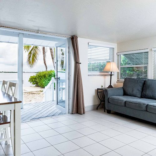 The image shows a bright living room with a gray sofa, a lamp, and a small dining table. It opens to a deck looking out over a beach with palm trees.