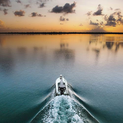 A boat is cruising on calm water towards the horizon during a picturesque sunset, creating a wake trail behind it under a sky adorned with clouds.