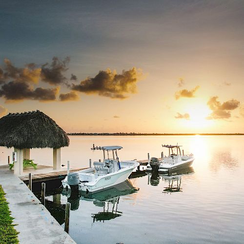 A serene sunset over calm water with boats docked by a thatched-roof hut and lush green grass, casting reflections on the water's surface.