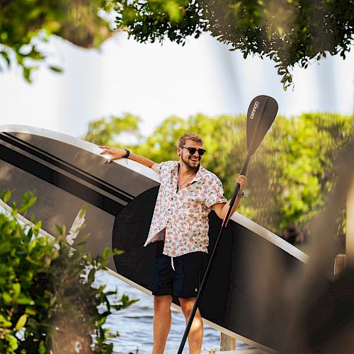 A person stands outdoors holding a paddleboard and paddle, surrounded by greenery. They are wearing shorts and a patterned shirt, and they are smiling.