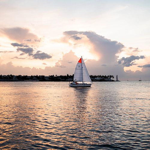 A sailboat glides on calm waters at sunset, with an island and dramatic clouds in the background. The sky is painted in soft hues of pink and orange.