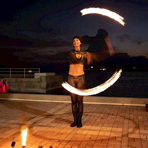 A person performs fire dancing at dusk, creating swirling light patterns with flames against a dark urban backdrop.