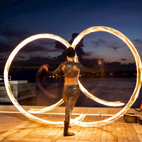 A performer is manipulating two large, illuminated hoops outdoors at dusk, creating bright, circular patterns against the dark sky.