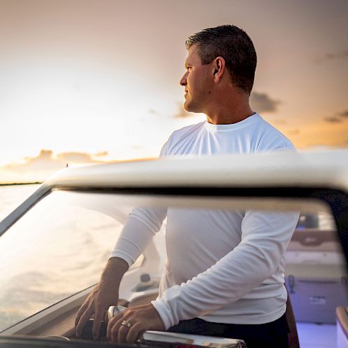 A man in a white shirt is steering a boat at sunset, looking off into the distance, with a calm sky in the background.