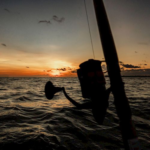 A fishing rod is silhouetted against a beautiful sunset over the sea, with gentle waves and an orange horizon casting a serene ambiance.