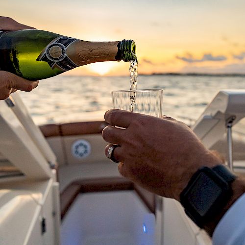 A bottle of champagne being poured into a glass on a boat, with a sunset over the water in the background, creating a serene and festive scene.