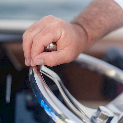 A person's hand is gripping the metallic steering wheel of a boat, with water visible in the background.