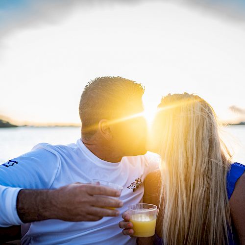 A couple is kissing on a boat during sunset. They each hold a drink, with the sun shining brightly in the background.