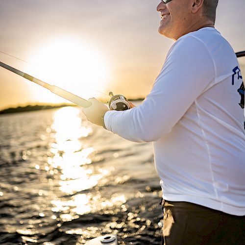 A person wearing sunglasses and a long-sleeve shirt is fishing on a boat as the sun sets over a body of water.