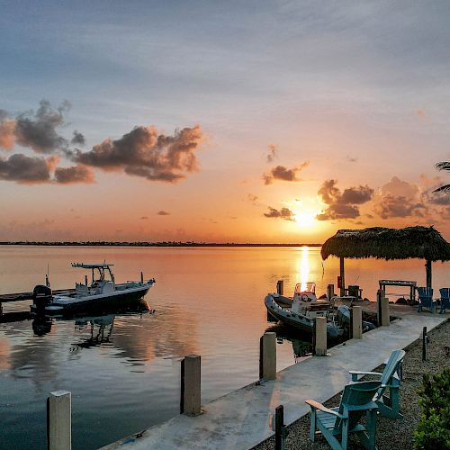 A tranquil sunset over a calm body of water with boats docked and a palm tree near a thatched hut in the foreground, ending the sentence.