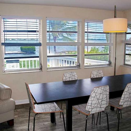 A dining area with a black table, six chairs, and a beige couch, located by windows offering a view of a peaceful waterfront scene.