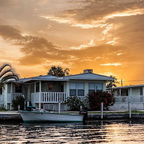 A serene sunset over a waterfront house with a boat docked nearby, palm trees, and warm, colorful skies reflecting off the water.