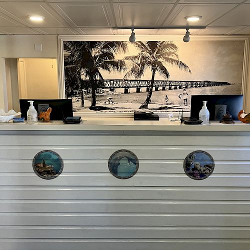 The image shows a white reception desk with three circular artwork discs. Behind it is a black and white photo of a beach scene with palm trees.