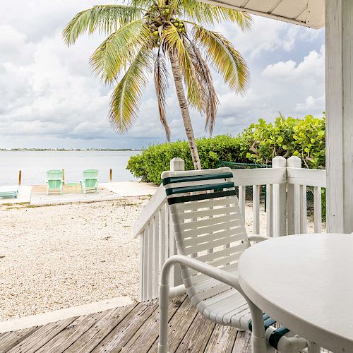 A cozy porch with white furniture overlooks a serene beach scene with palm trees and green lounge chairs near the water under a cloudy sky.