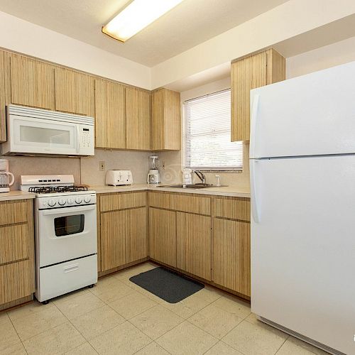 The image shows a compact kitchen with wooden cabinets, a white refrigerator, a small oven, a microwave, a sink, and various small appliances on the counter.