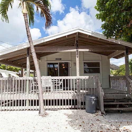 A quaint, single-story wooden cabin with a wraparound porch, palm trees, and a trash can in front, set against a partly cloudy sky.