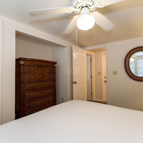 A bedroom with a white ceiling fan, a wooden dresser, a mirrored wall, and an open door, viewed from the bed. A round mirror and a nightstand are visible.